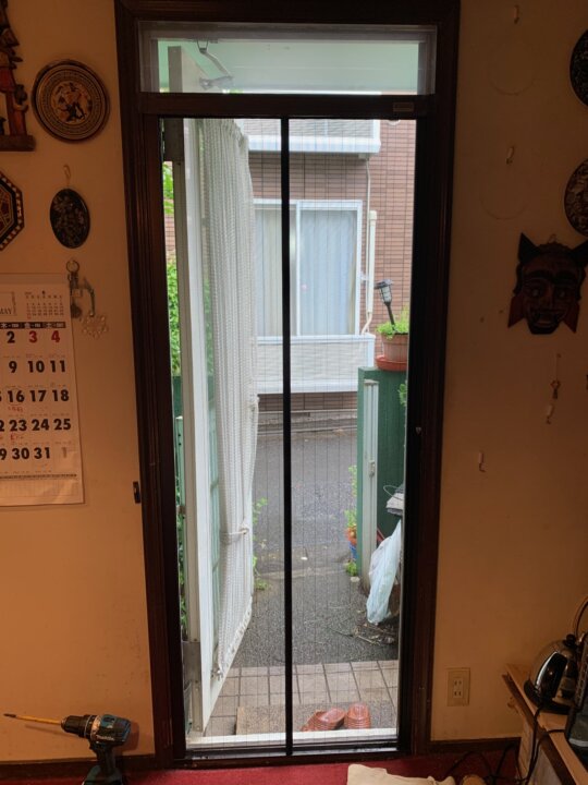 A view of a doorway with a newly installed pleated screen door, leading to a small outdoor area with plants. The interior shows a partially visible calendar and tools on the floor.